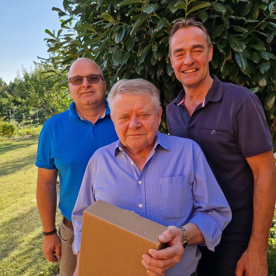 Drei Männer stehen zusammen in einem Garten und halten eine Schachtel. Der Mann in der Mitte ist älter und trägt ein lila Hemd. Der Mann links trägt ein blaues Hemd und der Mann rechts trägt ein dunkelblaues Polohemd.
