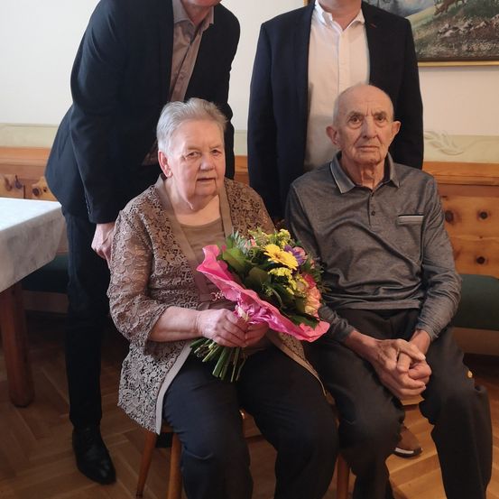 Zwei ältere Personen sitzen zusammen, eine hält einen Blumenstrauß. Hinter ihnen stehen zwei Männer in Anzügen, einer legt eine Hand auf die Schulter der sitzenden Frau. Im Hintergrund sind ein Tisch und eine Bank zu sehen.