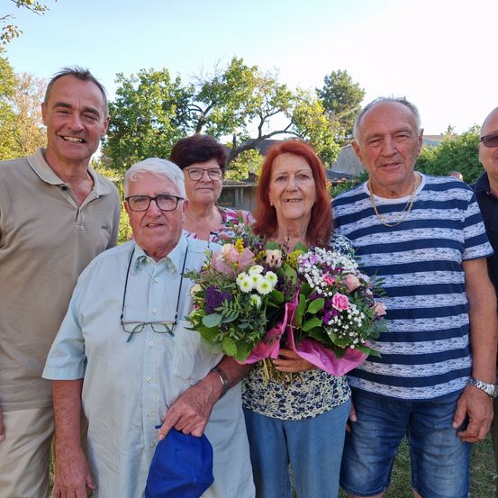 Eine Gruppe älterer Menschen steht draußen in einem Garten und lächelt für ein Foto. Die Frau in der Mitte hält einen Blumenstrauß. Hinter ihnen sind Bäume und ein Haus.