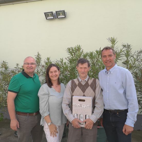 Vier Menschen stehen vor einer Wand mit Pflanzen. Der Mann auf der linken Seite trägt ein grünes Poloshirt. Die Frau in der Mitte trägt eine Halskette und eine Uhr. Der Mann in der Mitte hält eine Schachtel. Der Mann auf der rechten Seite trägt ein blaues Hemd.
