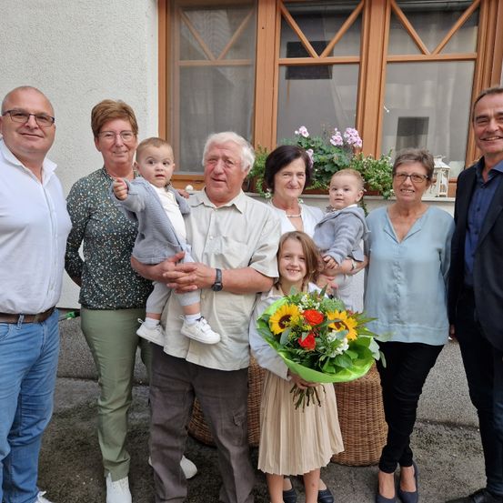 Ein Familienfoto zeigt eine Gruppe von Menschen vor einem Haus, wobei zwei Kinder von ihren Eltern gehalten werden und ein junges Mädchen einen Blumenstrauß hält.