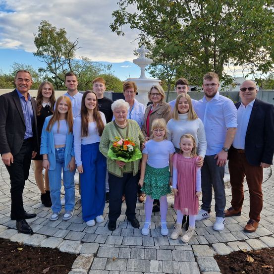 Eine Familie steht draußen mit einem Brunnen hinter ihnen. Eine Frau hält einen Blumenstrauß. Die Gruppe trägt eine Mischung aus lässiger und formeller Kleidung.