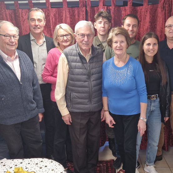 Eine Gruppe von Menschen, einschließlich eines älteren Paares, steht lächelnd für ein Foto vor einem roten Vorhang.