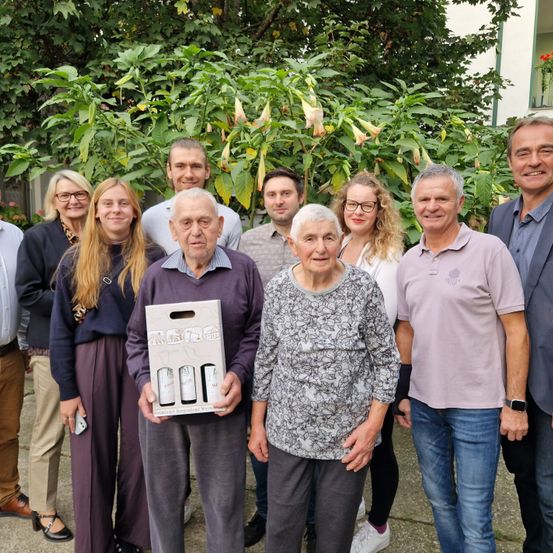 Eine Gruppe von Menschen steht in einem Garten, wobei ein Paar in der Mitte eine Weinschachtel hält. Dahinter ist eine große grüne Pflanze mit weißen Blüten zu sehen. Alle lächeln.