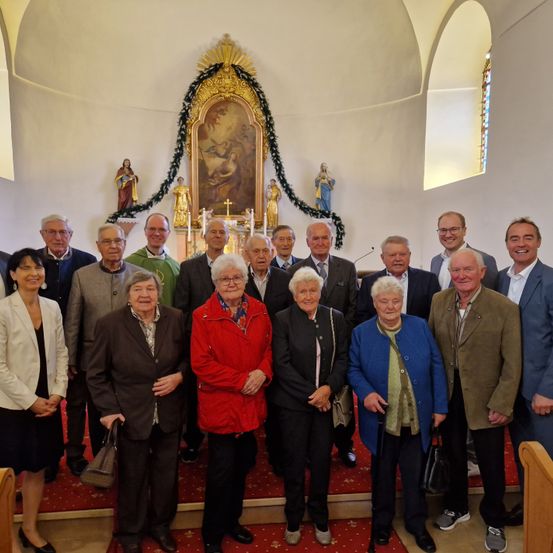 Eine Gruppe älterer Personen, sowohl Männer als auch Frauen, steht vor einem Kirchenaltar und lächelt für ein Gruppenfoto. Sie sind in Winterkleidung gekleidet.
