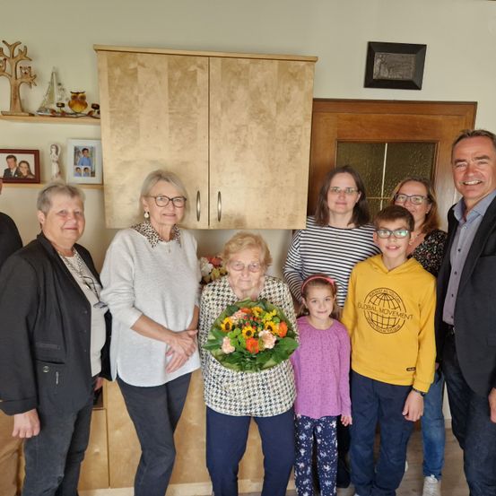 Eine Gruppe von Menschen, darunter eine ältere Frau mit einem Blumenstrauß, steht in einem Raum mit Holzschränken und einer Holztür.