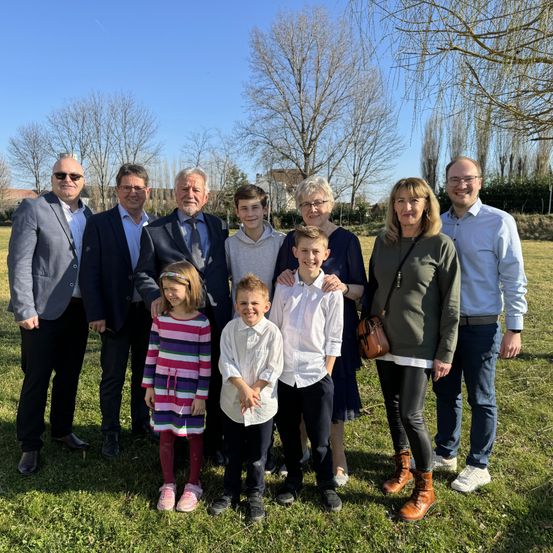 Eine Familie mit acht Personen steht auf einer grasbewachsenen Wiese, mit vier Erwachsenen und vier Kindern. Sie sind formell gekleidet, wobei zwei Frauen eine Brille tragen. Die Erwachsenen lächeln und die Kinder schauen in die Kamera.