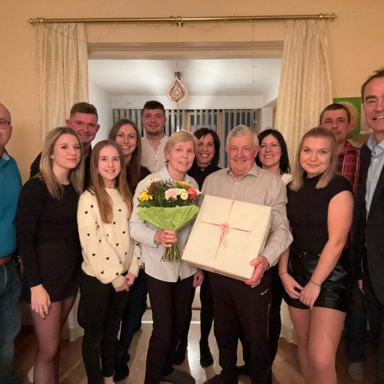 Eine Gruppe von Menschen steht drinnen und posiert für ein Foto. Eine Frau hält einen Blumenstrauß und eine Geschenkbox. Sie tragen Freizeitkleidung. Der Raum hat Vorhänge und Holzböden.