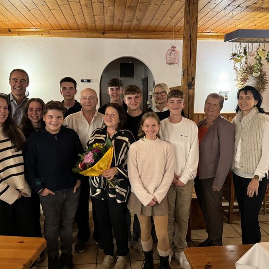 Eine Gruppe von Menschen steht in einem Raum mit Holzwänden und -decke. Sie lächeln und schauen zur Kamera. Einige halten Blumen. Hinter ihnen sind Tische und Stühle.