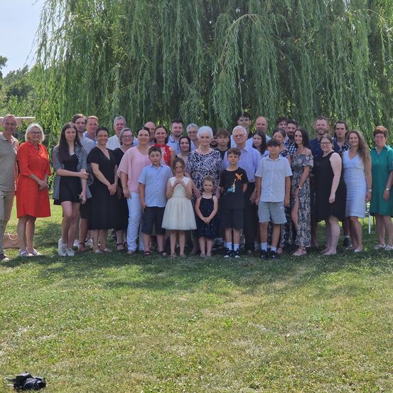 Eine große Gruppe von Menschen steht unter einer Weide und posiert für ein Foto. Verschiedene Kleidungsfarben sind sichtbar. Es gibt sowohl Erwachsene als auch Kinder.