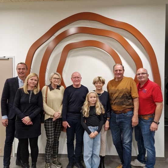 Eine Gruppe von Erwachsenen und einem jungen Mädchen posiert für ein Foto vor einem großen, welligen Wandkunstwerk. Die Erwachsenen sind in verschiedenen Outfits gekleidet, und das Mädchen trägt ein schwarzes Shirt und Jeans. Sie alle scheinen zu lächeln.