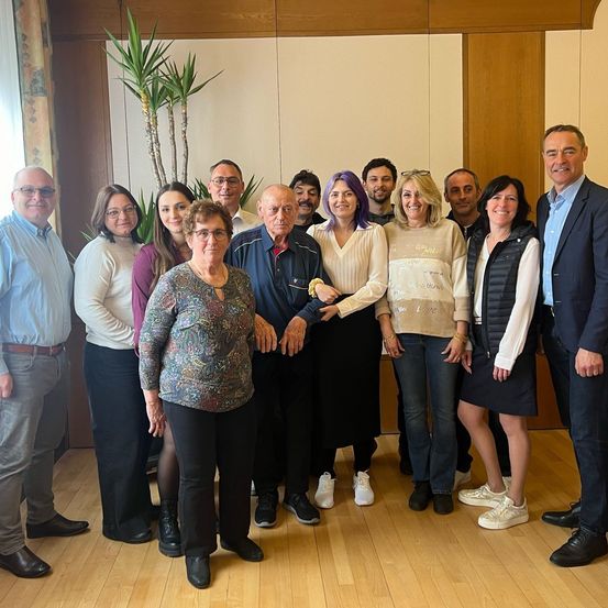 Eine Gruppe von Personen, darunter Männer und Frauen, posiert für ein Foto in einer Innenumgebung. Sie stehen vor einer Holzwand und einer großen Pflanze.