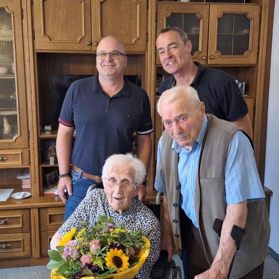 Drei Männer und eine ältere Frau im Rollstuhl halten Blumen in einem Raum mit Holzschränken.