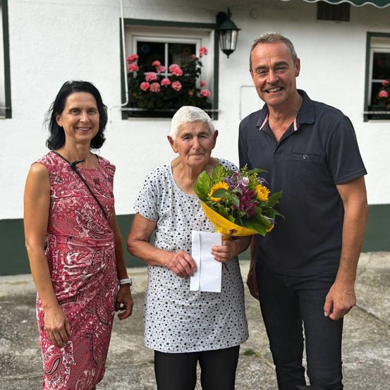 Drei Menschen stehen vor einem weißen Gebäude mit Blumen im Fenster. Die Frau auf der linken Seite lächelt und trägt ein Kleid. Die Frau in der Mitte hält einen Blumenstrauß und ein Stück Papier. Der Mann auf der rechten Seite lächelt und trägt ein Poloshirt.