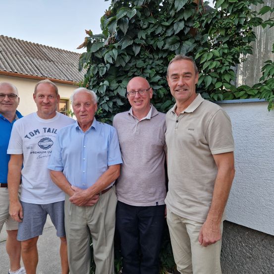 Fünf Männer stehen lächelnd im Freien. Einer trägt ein blaues Shirt, einer ein weißes, einer ein graues und zwei beigefarbene Shirts. Dahinter ein grüner Busch und eine weiße Wand.