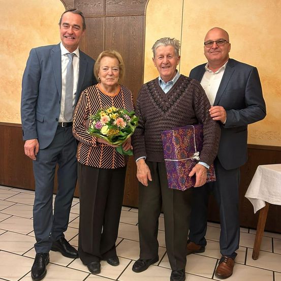 Vier Personen stehen zusammen, lächeln und scheinen für ein Foto zu posieren. Eine Frau hält einen Blumenstrauß, während ein Mann ein eingewickeltes Geschenk hält.