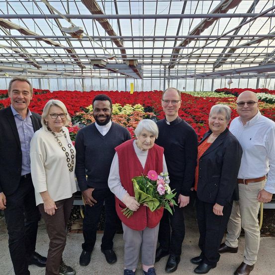 Sieben Erwachsene stehen in einem Gewächshaus mit rotem Blumenhintergrund. Eine Frau hält einen Blumenstrauß.