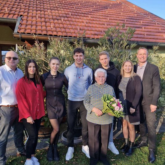 Eine Gruppe von Menschen steht vor einem Haus mit einem Ziegeldach. Sie lächeln und posieren für ein Foto. Eine Frau in der Mitte hält einen Blumenstrauß.