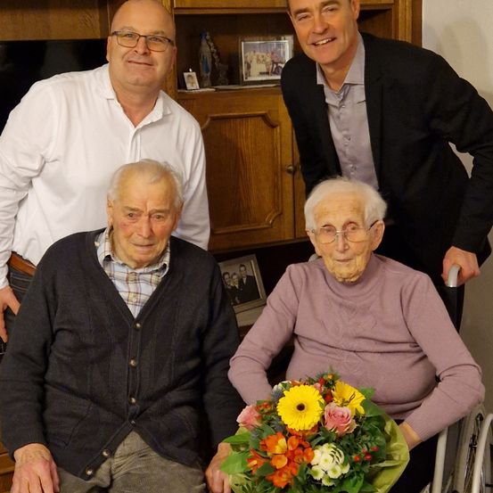 Drei ältere Erwachsene lächeln gemeinsam, einer im Rollstuhl mit Blumen, ein anderer mit einem Blumenstrauß, und zwei stehen dahinter. Sie befinden sich in einem Raum mit Holzschränken und gerahmten Fotos.