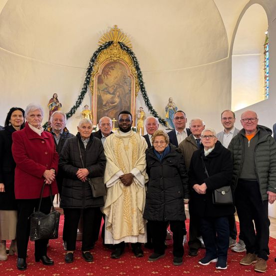 Eine Gruppe von Menschen in warmer Winterkleidung steht in einer Kirche und posiert für ein Foto mit einem Priester. Hinter ihnen ein Altar mit einem Gemälde und Dekorationen.