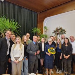 Eine Gruppe von Menschen ist für ein Foto mit einer lächelnden älteren Frau mit Blumen versammelt. Sie stehen in der Nähe von Pflanzen und einer weißen Wand mit einer Blumenarrangement. Der Mann im Anzug lächelt und die Frau in Weiß lächelt.
