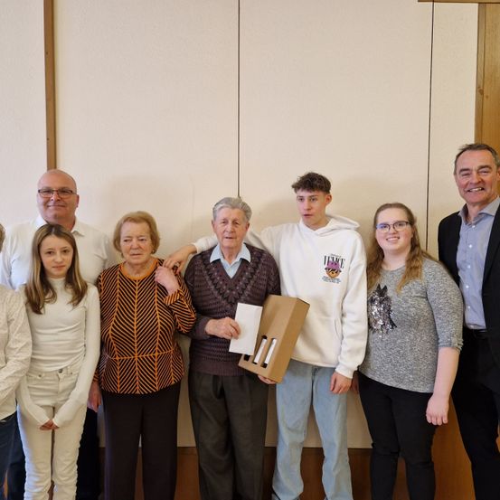 Eine Gruppe von Menschen steht lächelnd vor einer hellen Wand. Ein älterer Mann hält ein Papier, und eine Frau in einem gemusterten Shirt steht neben ihm. Der Mann rechts trägt einen Anzug und eine Krawatte.