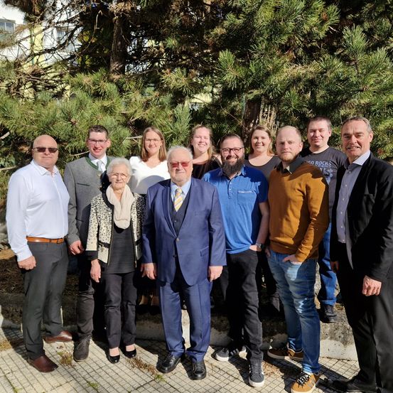 Eine Gruppe von Personen, möglicherweise ein Team oder eine Familie, steht vor einer Kiefer und lächelt für ein Foto.