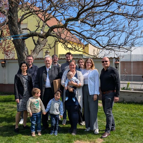 Ein Familienfoto, aufgenommen in einem Garten mit einem gelben Haus im Hintergrund. Mehrere Erwachsene und Kinder posieren für das Bild.