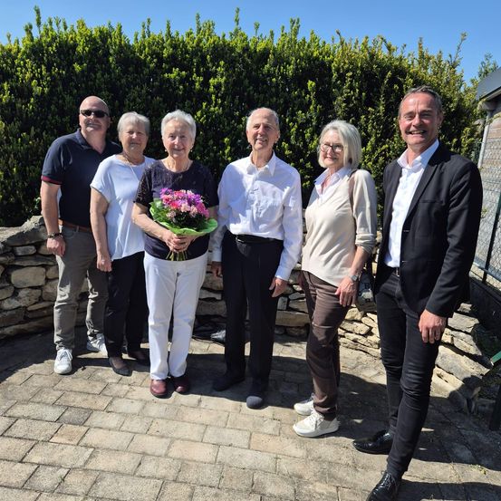 Eine Gruppe von sechs Menschen steht auf einem Ziegelweg und lächelt für ein Foto. Eine Frau hält einen Blumenstrauß. Hinter ihnen steht eine Steinmauer.