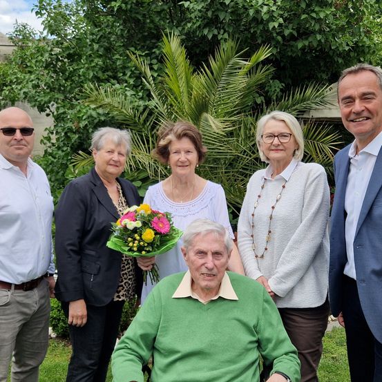 Eine Gruppe von fünf Personen steht in einem Garten, wobei ein älterer Mann im Rollstuhl einen Blumenstrauß hält. Hinter ihm stehen zwei Frauen und zwei Männer. Bäume und Pflanzen sind im Hintergrund.