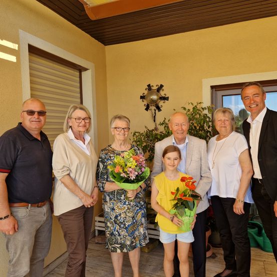 Sieben Menschen, darunter ein junges Mädchen mit Blumen, stehen auf einer Veranda mit gelben Wänden. Sie scheinen für ein Foto zu posieren.