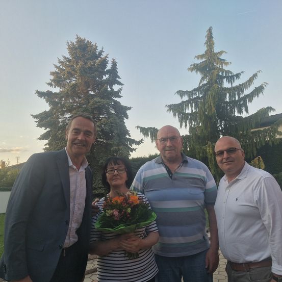 Vier Menschen stehen lächelnd vor Bäumen. Die Frau hält einen Blumenstrauß. Die Männer tragen Brillen und Anzüge. Der Himmel ist klar.