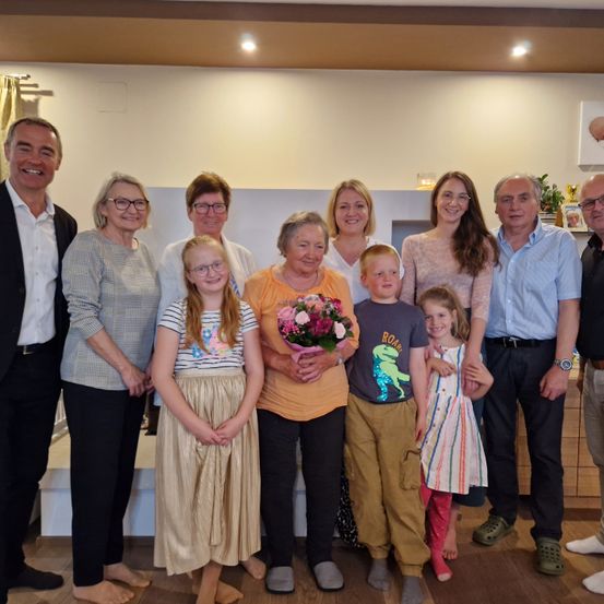 Eine Familiengruppe posiert für ein Foto in einem Raum mit Holzboden. Es gibt zwei junge Mädchen, Erwachsene und Kinder. Die Frau in der Mitte hält einen Blumenstrauß.