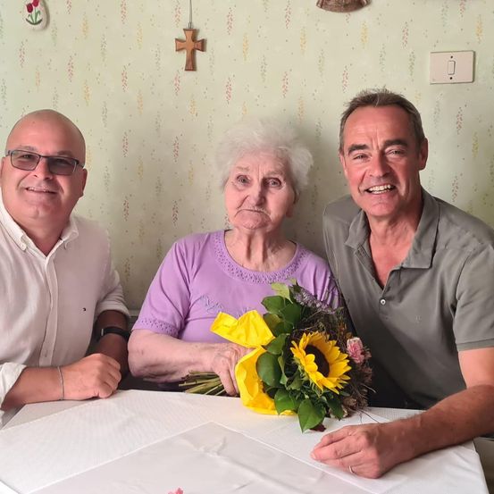 Drei Menschen lächeln in die Kamera. Eine ältere Frau hält einen Blumenstrauß. Ein Mann in einem grauen Hemd ist neben ihr, und ein anderer Mann in einem weißen Hemd ist auf ihrer linken Seite. Die Wand hinter ihnen hat ein Kreuz und einen Lichtschalter.