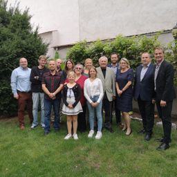 Eine Gruppe von Menschen steht in einem Garten, schick gekleidet, mit einem Gebäude und Grünpflanzen im Hintergrund.