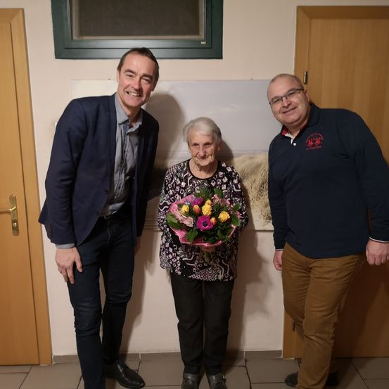Drei Menschen stehen lächelnd vor einer Wand mit einem Gemälde. Die Frau hält einen Blumenstrauß in der Hand.