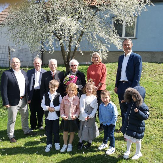 Eine Gruppe von Menschen, möglicherweise eine Familie, steht auf einer Rasenfläche. Sie lächeln und scheinen für ein Foto zu posieren. Eine Frau hält Blumen. Vor ihnen steht ein Baum. Dahinter befindet sich ein Haus mit einem Zaun.