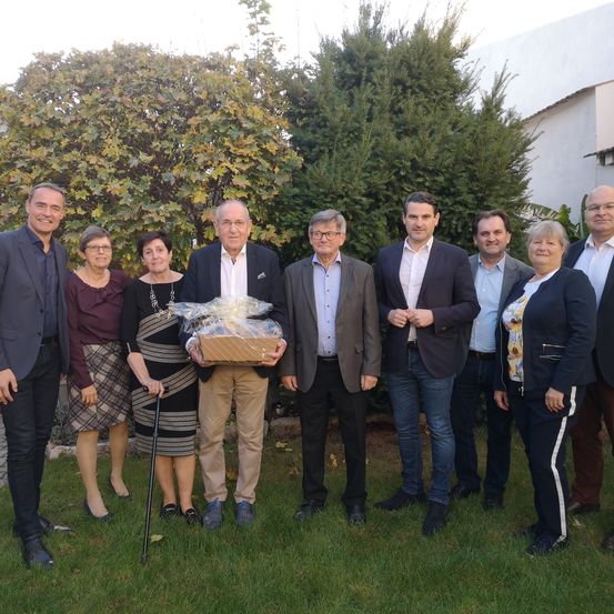 Eine Gruppe von Menschen steht auf einem Rasen und lächelt für ein Foto. Ein Mann in der Mitte hält ein verpacktes Geschenk. Sie sind umgeben von Bäumen und einem weißen Gebäude.