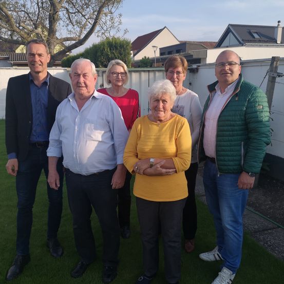 Sechs Menschen stehen auf einem grünen Rasen, lächeln und posieren für ein Foto. Hinter ihnen sind Häuser und Bäume zu sehen.