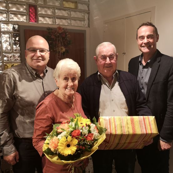 Vier Menschen stehen in einem Raum und lächeln. Eine ältere Frau hält einen Blumenstrauß und eine Geschenkbox. Der Mann auf der rechten Seite trägt einen schwarzen Anzug und der Mann auf der linken Seite trägt eine Brille.