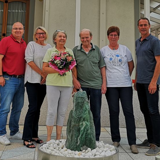 Fünf Erwachsene, zwei Männer und drei Frauen, stehen vor einem Gebäude. Eine Frau in der Mitte hält einen Blumenstrauß. Sie alle lächeln.