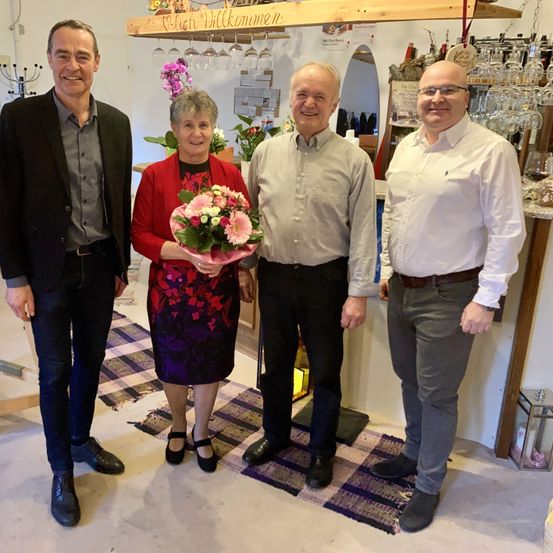 Vier Menschen stehen in einem Raum und lächeln. Die Frau hält einen Blumenstrauß. Hinter ihnen steht ein Schild mit der Aufschrift 'Frisch Willkommen'. An der Wand sind Regale mit Weingläsern und Flaschen angebracht.