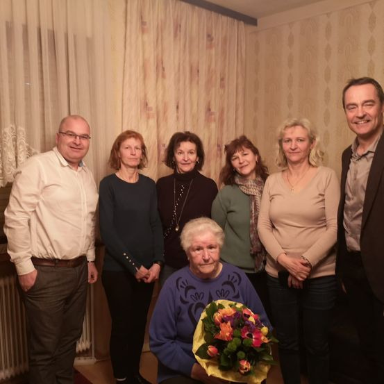 Eine Gruppe Erwachsener, darunter eine ältere Frau mit Blumen, steht in einem Raum mit gemusterter Tapete und weißen Vorhängen.