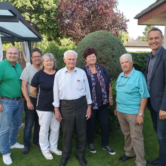 Eine Gruppe Erwachsener steht draußen in einem Garten. Sie alle lächeln und scheinen für ein Foto zu posieren. Hinter ihnen befinden sich Hecken und ein Haus.