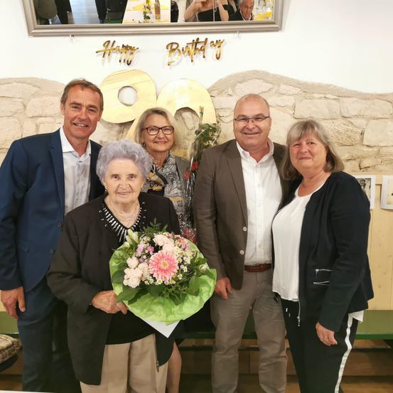 Eine Gruppe von Menschen steht zusammen und feiert einen Geburtstag. Eine ältere Frau hält einen Blumenstrauß. Hinter ihr stehen zwei Frauen und zwei Männer, die lächeln. Über ihnen ist ein Geburtstagsbanner zu sehen.