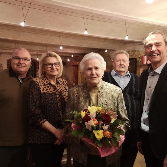 Fünf Personen sind drinnen versammelt und lächeln für ein Foto. Eine ältere Frau in der Mitte hält einen Blumenstrauß. Hinter ihr steht ein Mann mit Brille neben einer Frau in einer Leoparden-Bluse. Ein weiterer älterer Mann steht hinter ihnen, und ein Mann in einem Anzug steht rechts.