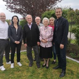 Eine Gruppe von Menschen, darunter eine Frau in einer rosa Bluse und ein Mann in einem schwarzen Anzug, posiert für ein Foto im Garten.
