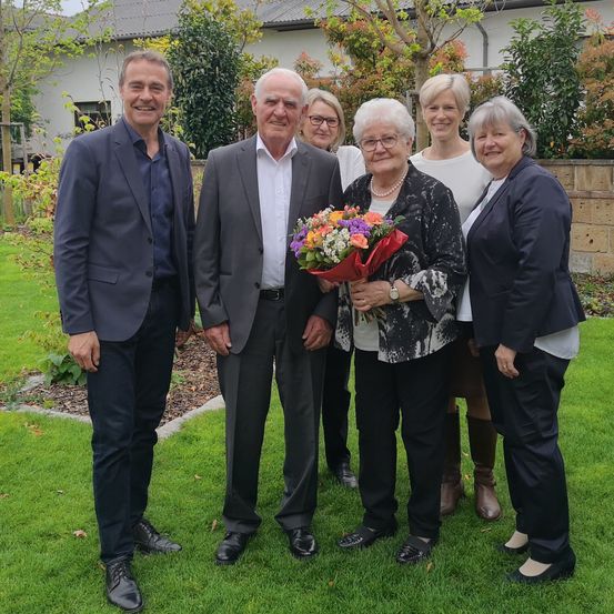 Eine Gruppe von Menschen steht in einem Garten und lächelt für ein Foto. Eine Frau hält einen Blumenstrauß und hinter ihr tragen ein Mann und eine Frau formelle Kleidung. Sie sind von Pflanzen umgeben und im Hintergrund befindet sich ein Gebäude.