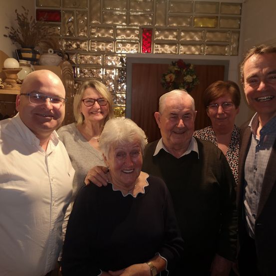 Sechs Menschen stehen lächelnd in einem Raum mit einer Mosaikwand und einem Kranz. Ein Mann und eine Frau sind an den gegenüberliegenden Seiten.