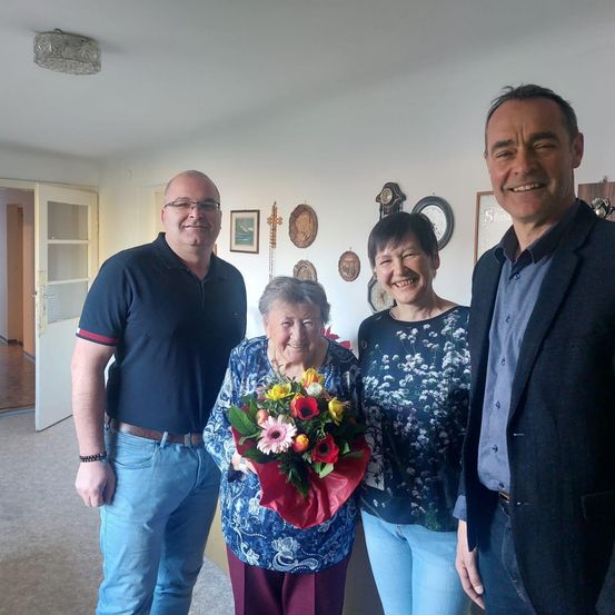 Drei Menschen stehen in einem Raum mit einer lächelnden älteren Frau, die Blumen hält. Ein Mann links und rechts lächelt. Eine Frau in der Mitte hat ein Blumendesign auf ihrem Oberteil.